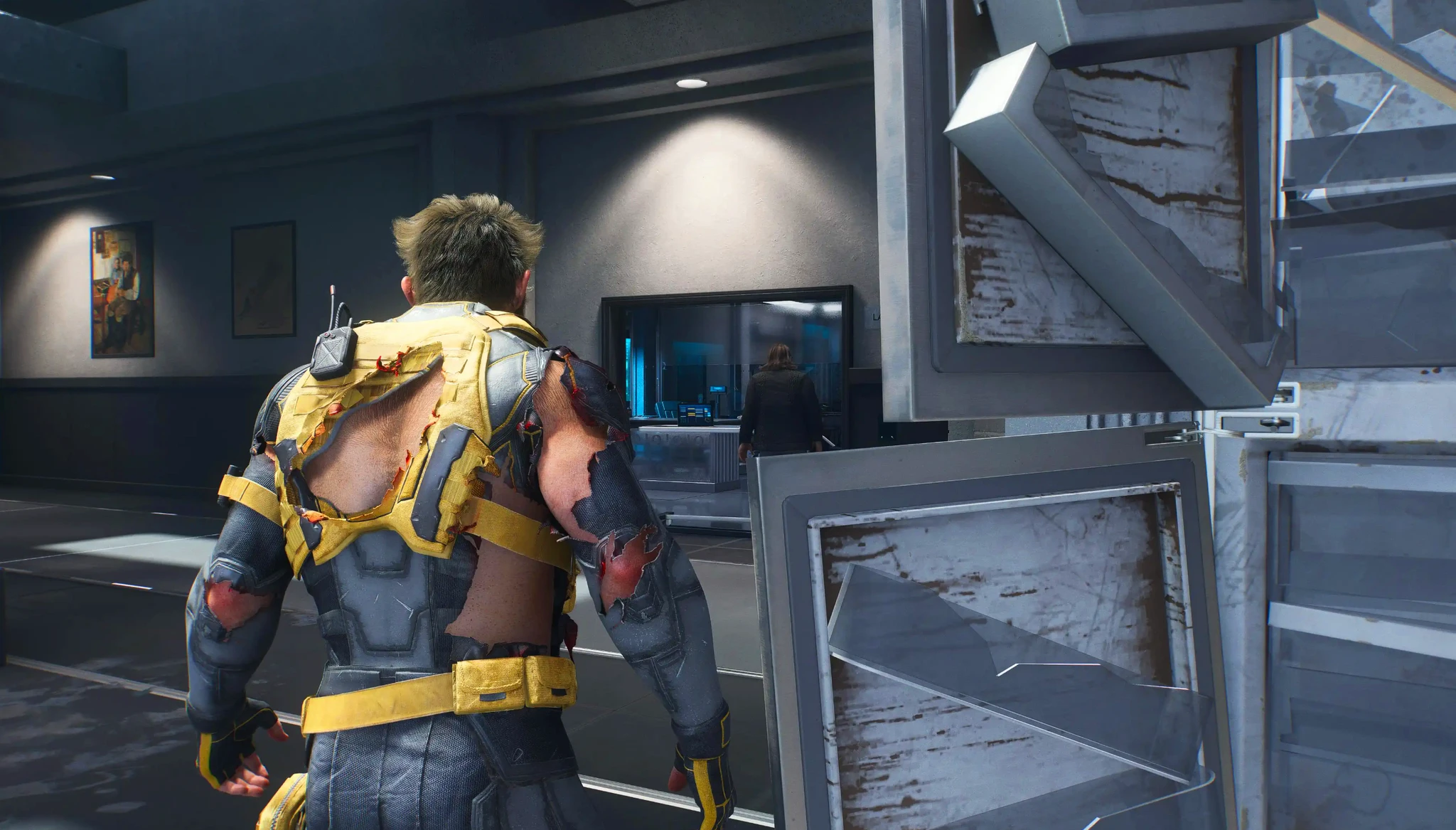 Wolverine i trasig gul och grå dräkt går genom en mörk korridor i Marvel's Wolverine, med söndersmulade metallskåp i förgrunden och en skärm i bakgrunden.
