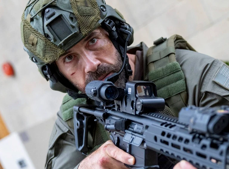 Skäggig soldat i hjälm och skyddsväst siktar med ett automatkarbin med kikarsikte, med smutsigt ansikte och intensiv blick.