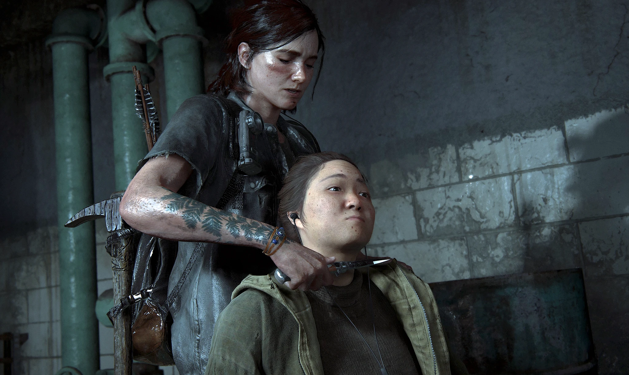 Ellie med tatuerad underarm står bakom en sittande kvinna i grön jacka och håller en kniv mot hennes hals i en dunkel, kaklad källare i The Last of Us Part II.
