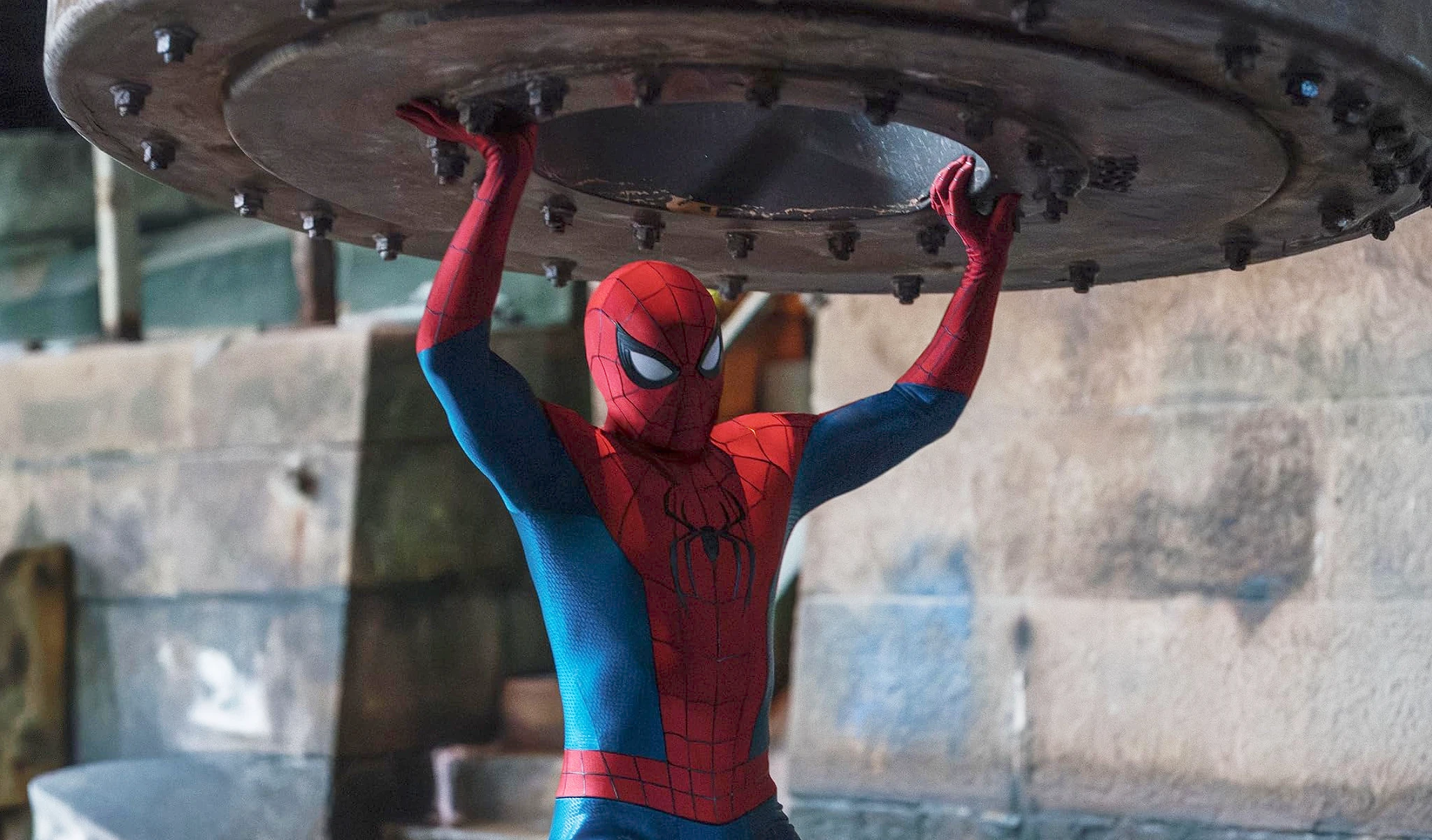 Spider-Man i rödblå dräkt står i en gammal industrilokal och lyfter med sträckta armar en enorm rund metallplatta med bultar över huvudet.