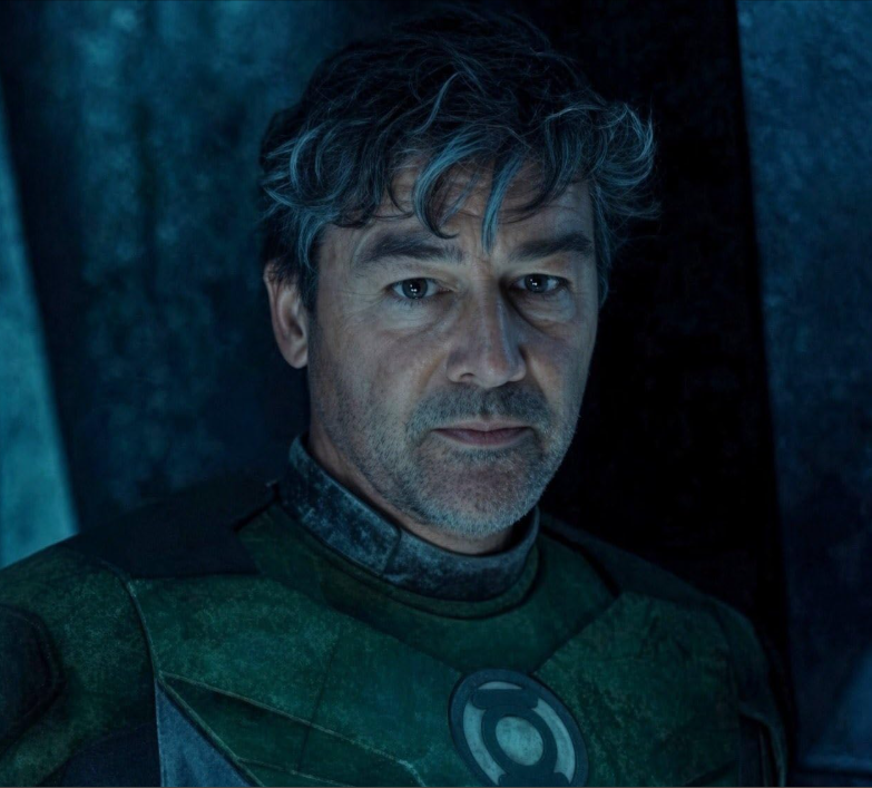 Närbild på en medelålders man med rufsigt grått hår och skäggstubb i en grön rustning med Green Lantern-emblem på bröstet, i dunkelt blåaktigt ljus.