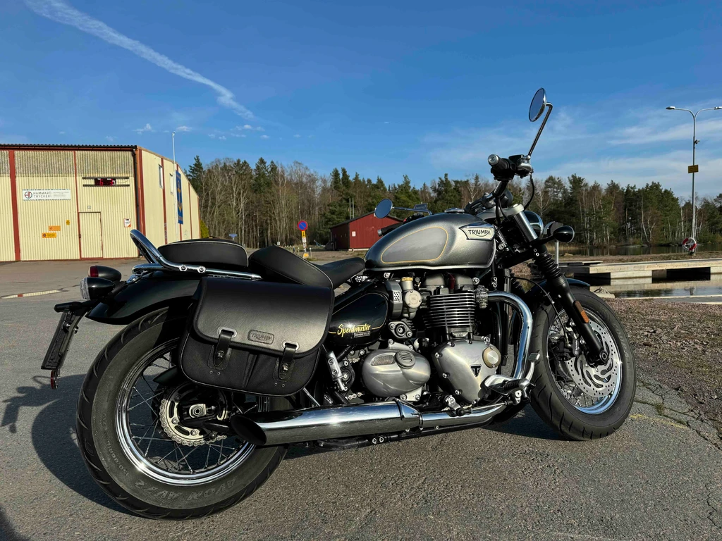 Grå och svart Triumph Bonneville Speedmaster med kromade avgasrör och svarta sadelväskor, parkerad på asfalt en solig dag med blå himmel och skog bakom.