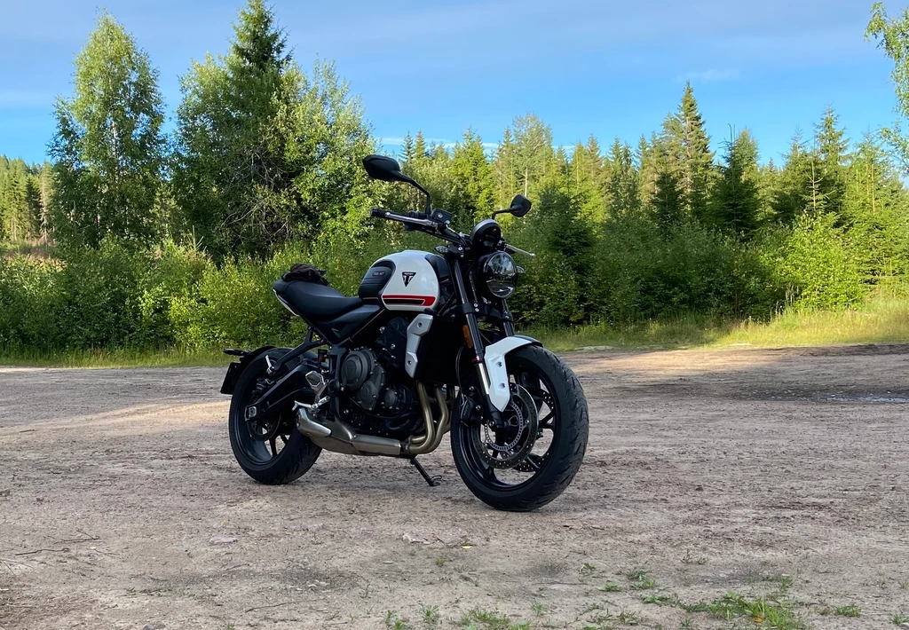Vit och svart Triumph Trident 660 står på en grusplan framför grönskande barrskog i kvällssol under blå himmel.