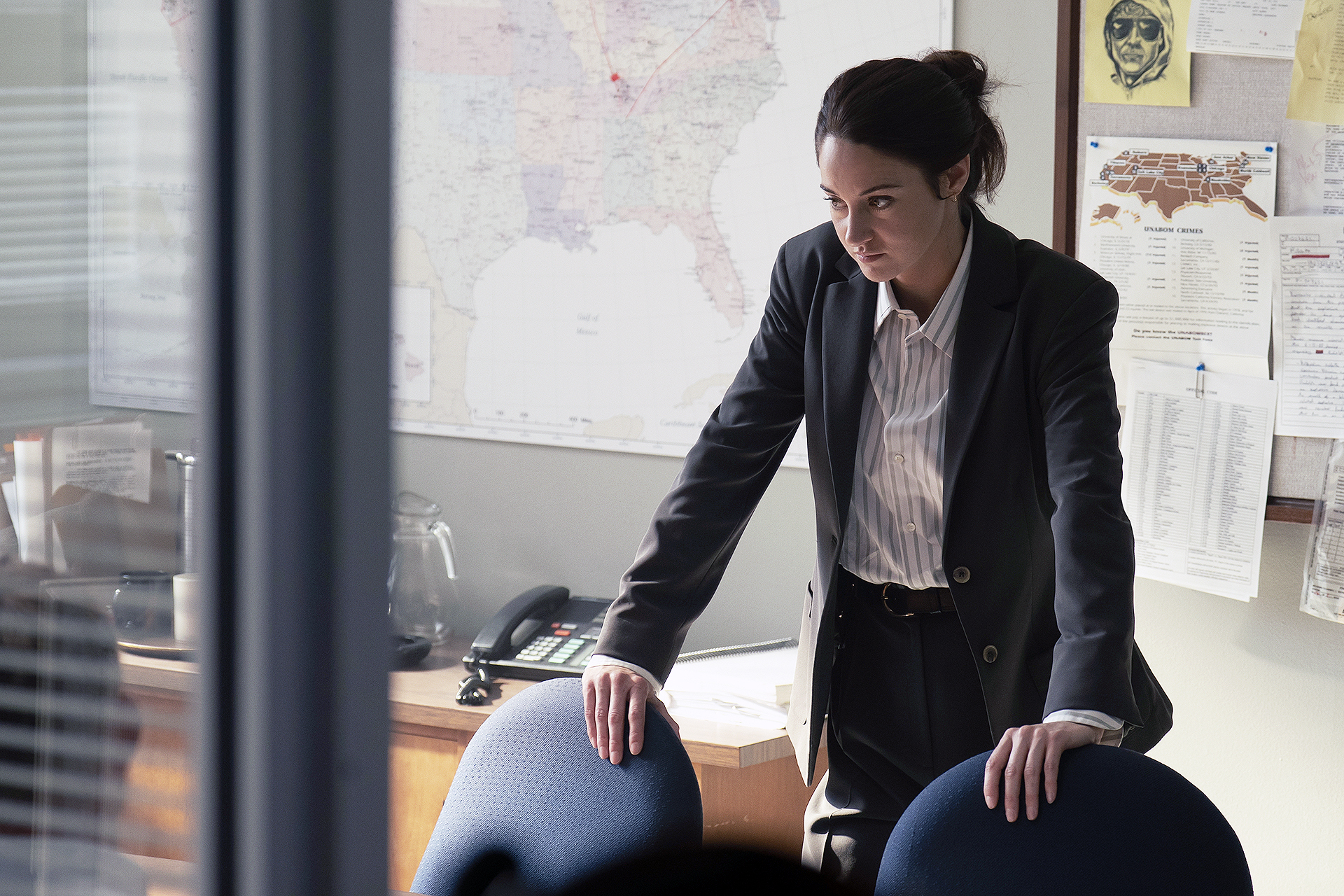"A woman in a suit leans on a conference table in an office, appearing serious and focused. The background features a large map, bulletin board with notes, a desk phone, and documents, suggesting an investigative or professional setting."