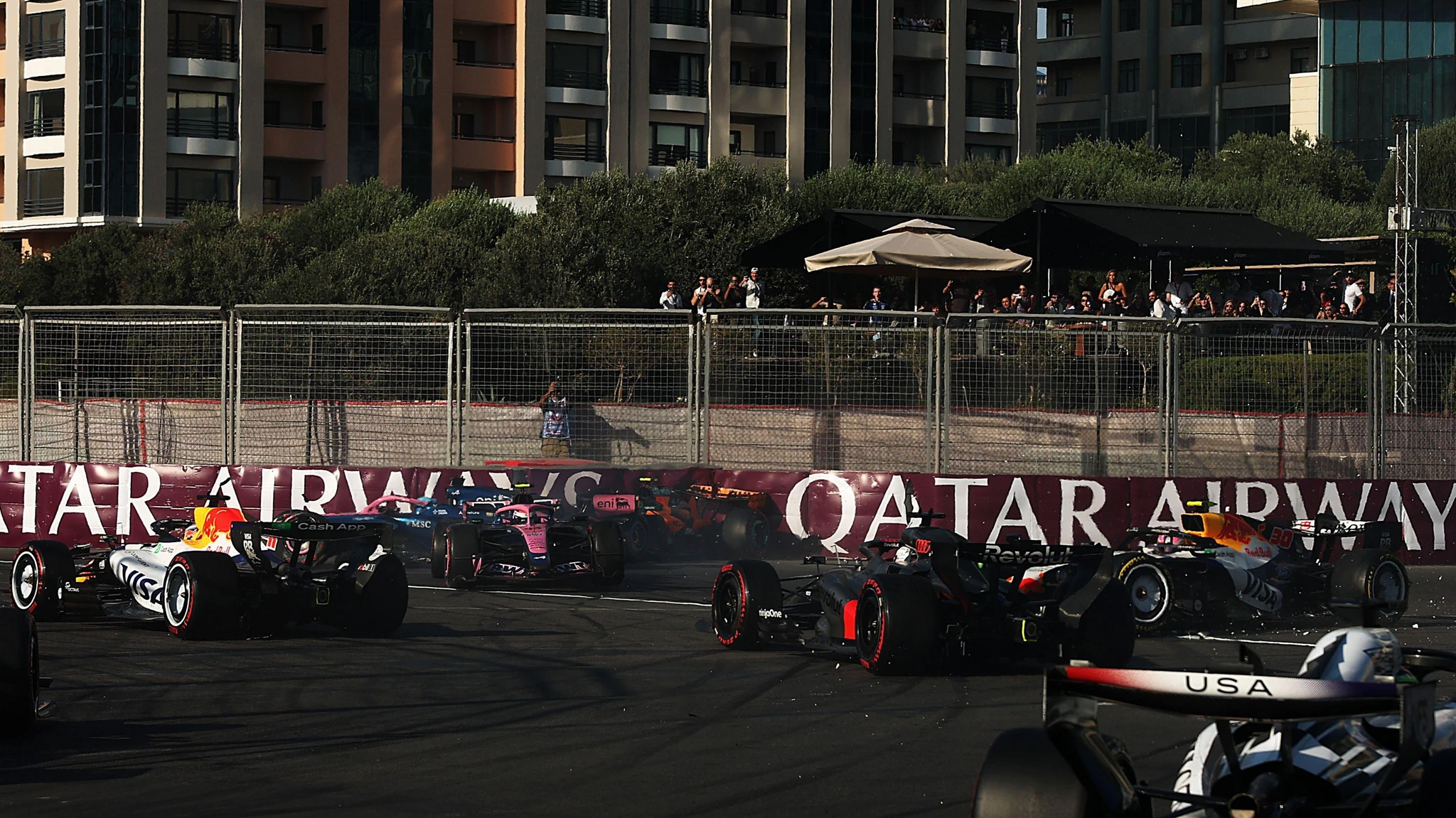 Flera Formel 1-bilar trängs och krockar på banan framför en Qatar Airways-skylt, med åskådare bakom stängslet och höghus i bakgrunden.