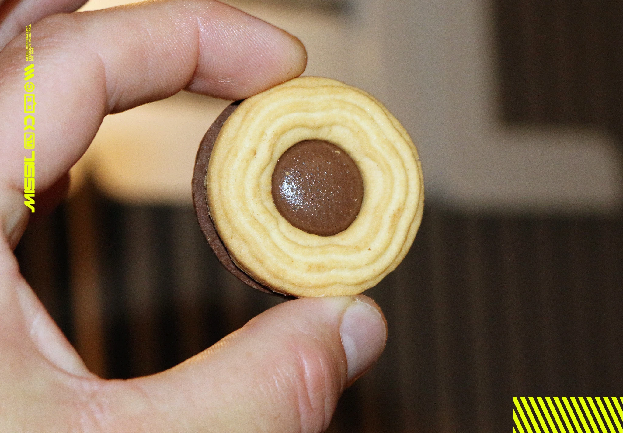 En hand håller upp en rund kaka med ljus, räfflad yta och en chokladklick i mitten. Undersidan är av mörk choklad.
