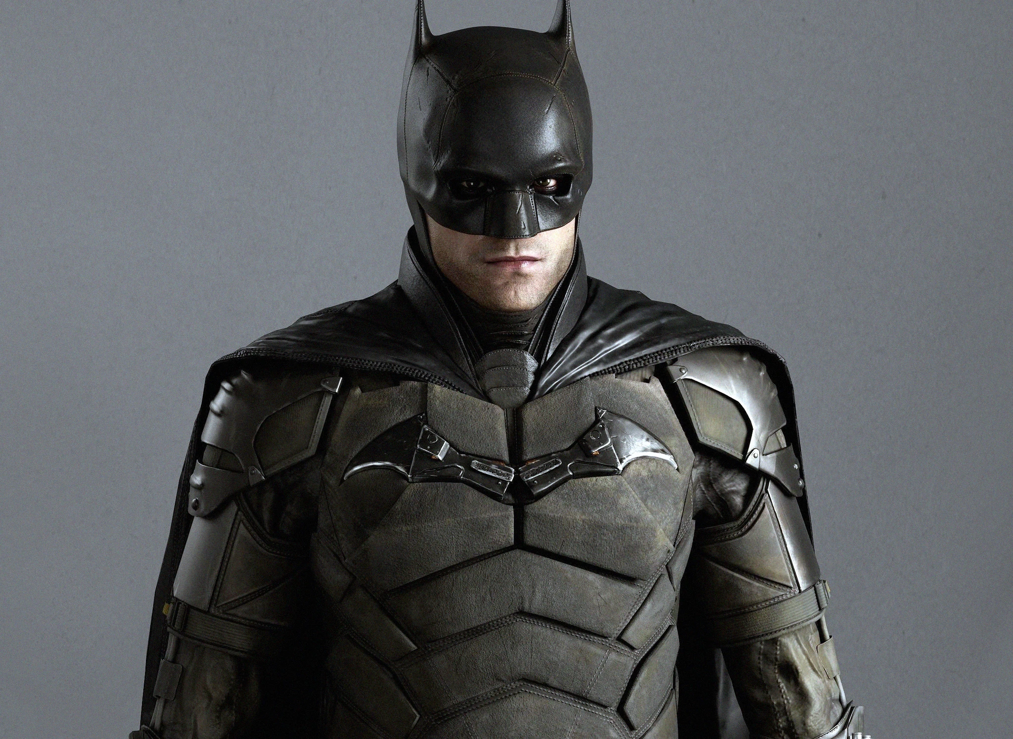 Batman i mörk, sliten pansardräkt med cape och spetsig mask framför en grå bakgrund, med flaggermusemblem på bröstet och blicken rakt fram.