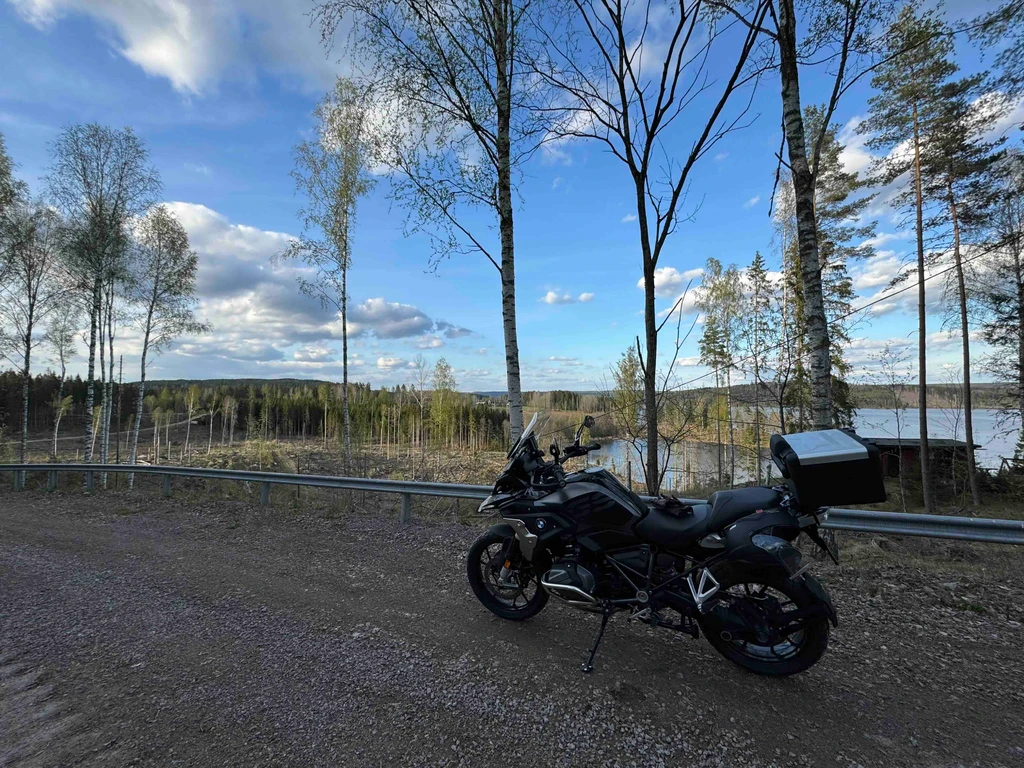 Svart äventyrsmotorcykel med koffert står parkerad på en grusväg bredvid ett vägräcke, med björkar, skog och en sjö i bakgrunden under blå himmel.