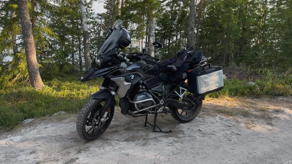 Svart BMW-motorcykel av äventyrsmodell med sidokappor och bagage står uppställd på stödet på en sandig skogsplats i kvällssol.