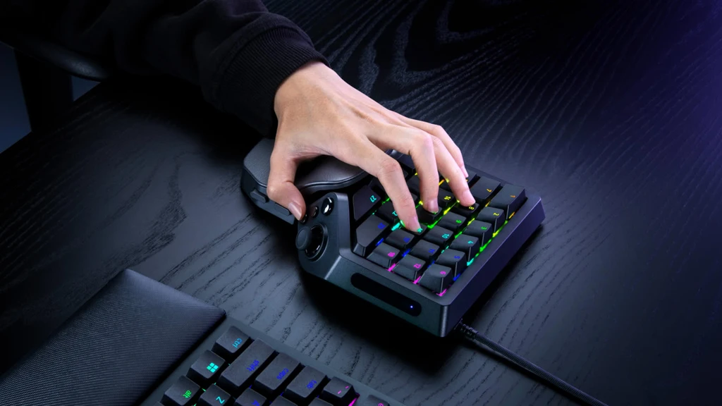 En hand vilar på Razer Tartarus V2 Pro, ett svart enhandstangentbord med färgglatt RGB-ljus under tangenterna, på ett mörkt träskrivbord bredvid ett vanligt tangentbord.
