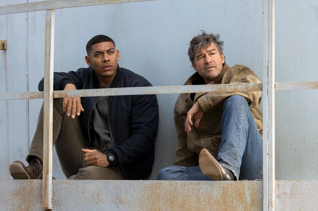 Aaron Pierre som John Stewart och Kyle Chandler som Hal Jordan i Lanterns sitter bredvid varandra bakom ett rostigt räcke och blickar allvarligt åt sidan.