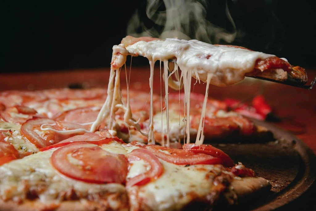 En pizzaskiva lyfts med spatel från en ångande pizza med tomatskivor, och den smälta osten dras ut i långa trådar.