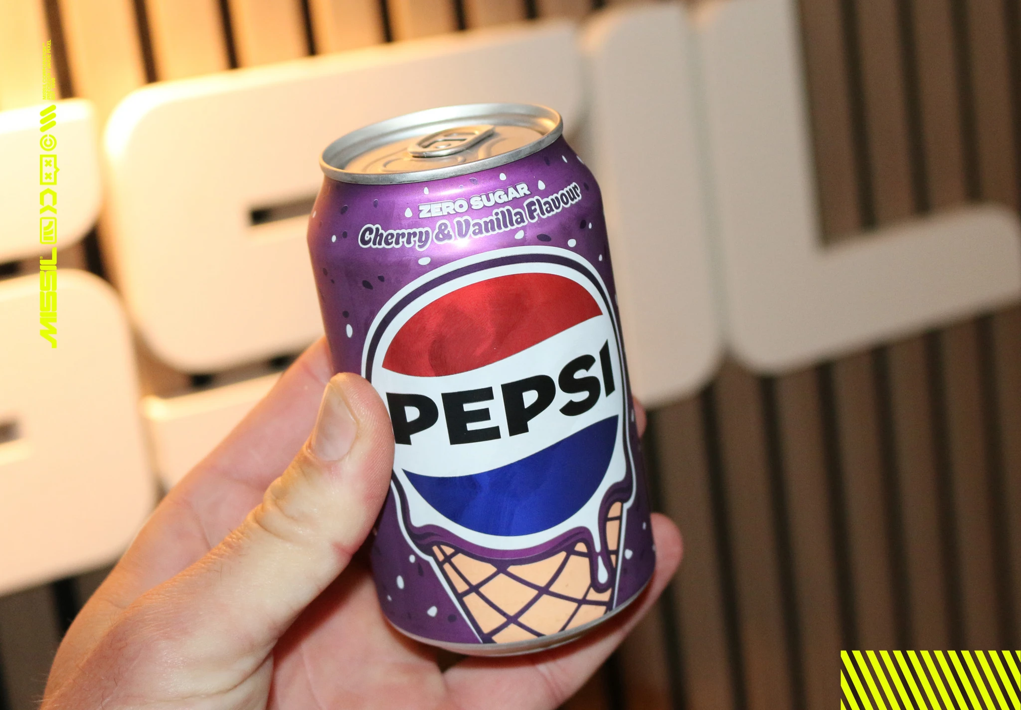 En hand håller upp en lila burk Pepsi Zero Sugar med smak av körsbär och vanilj, med en glassstrut på burken, framför vita bokstäver på en ribbad vägg.