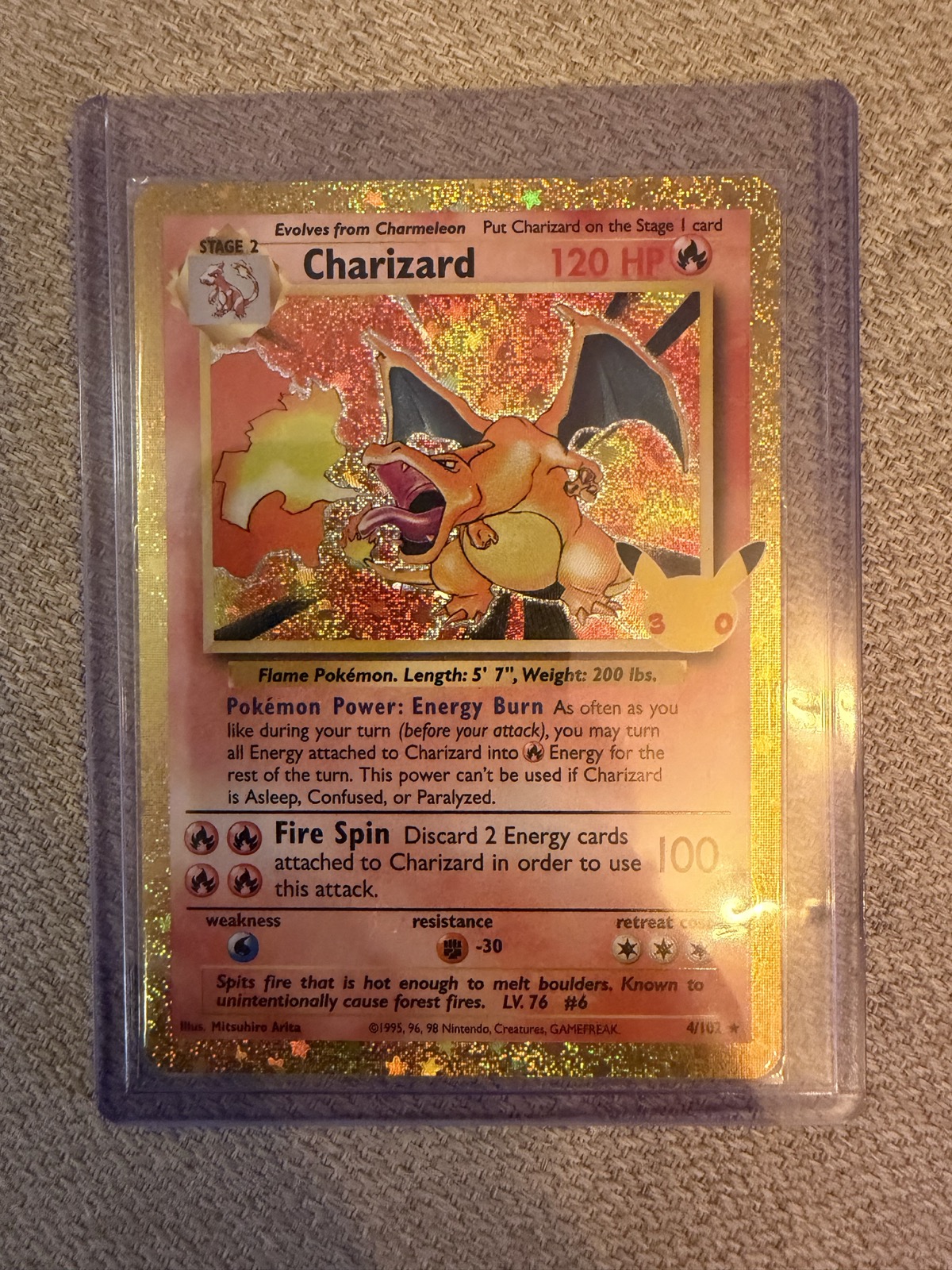 Glittrande Pokémon-kort med Charizard, 120 HP, i ett skyddande plastfodral. Kortet har guldram och 30-årsjubileumsmärke, och Charizard spyr eld.