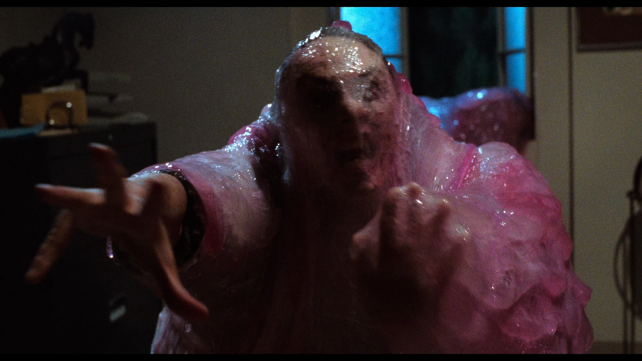 En person som delvis är uppslukad av en rosa, geléartad massa sträcker ut handen mot kameran i ett dunkelt rum, en scen ur skräckfilmen The Blob från 1988.
