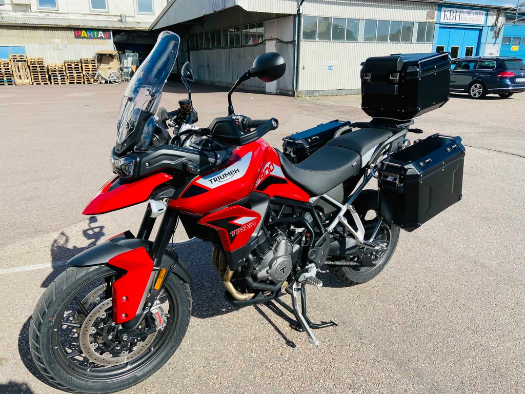 Röd Triumph Tiger 900 med svarta aluminiumväskor och toppbox, parkerad på sidstödet på ett asfalterat industriområde en solig dag.