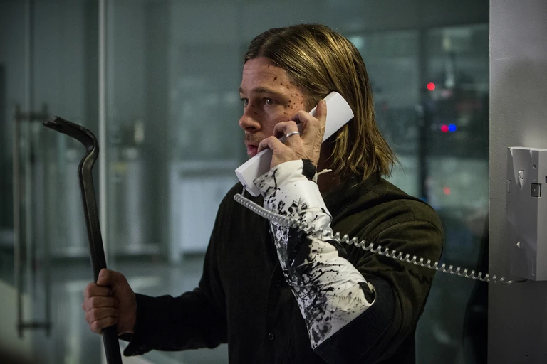Brad Pitt som Gerry Lane i World War Z talar i en väggtelefon med kråkfot i ena handen. Hans ärm är nedstänkt med mörk smuts eller blod.