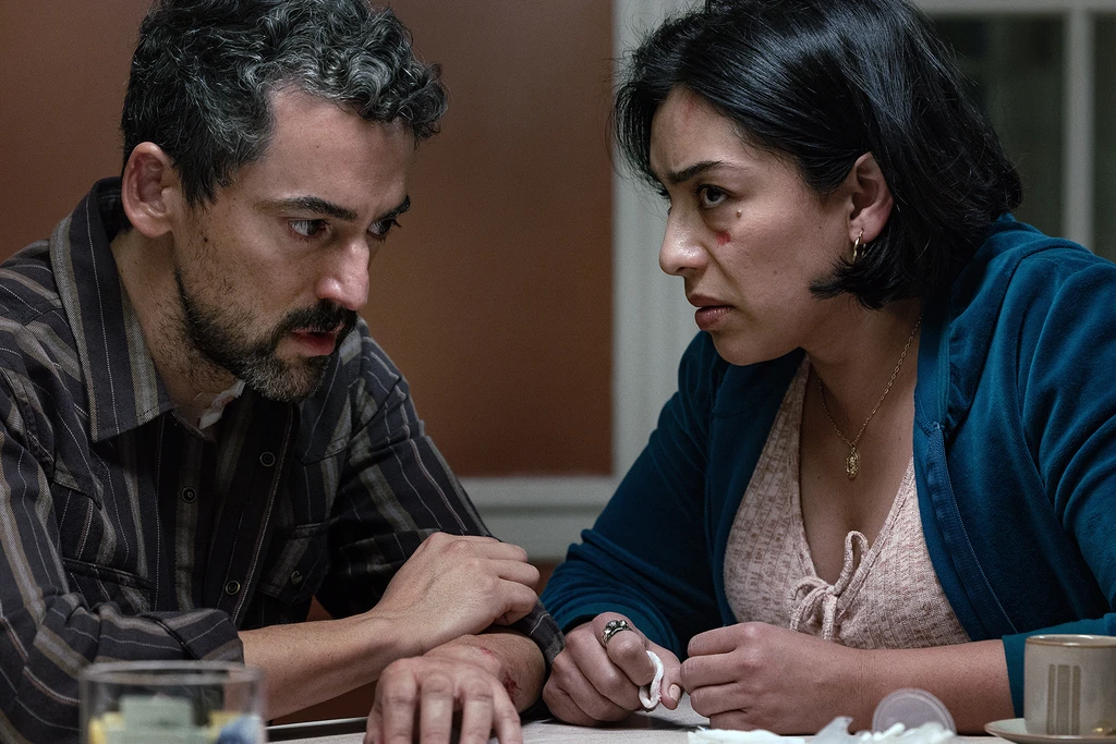 "Two concerned adults sit closely at a table in a dimly lit room, engaged in a serious conversation. The setting is a domestic environment with a warm-toned wall and household items present, suggesting an intense or emotional moment."