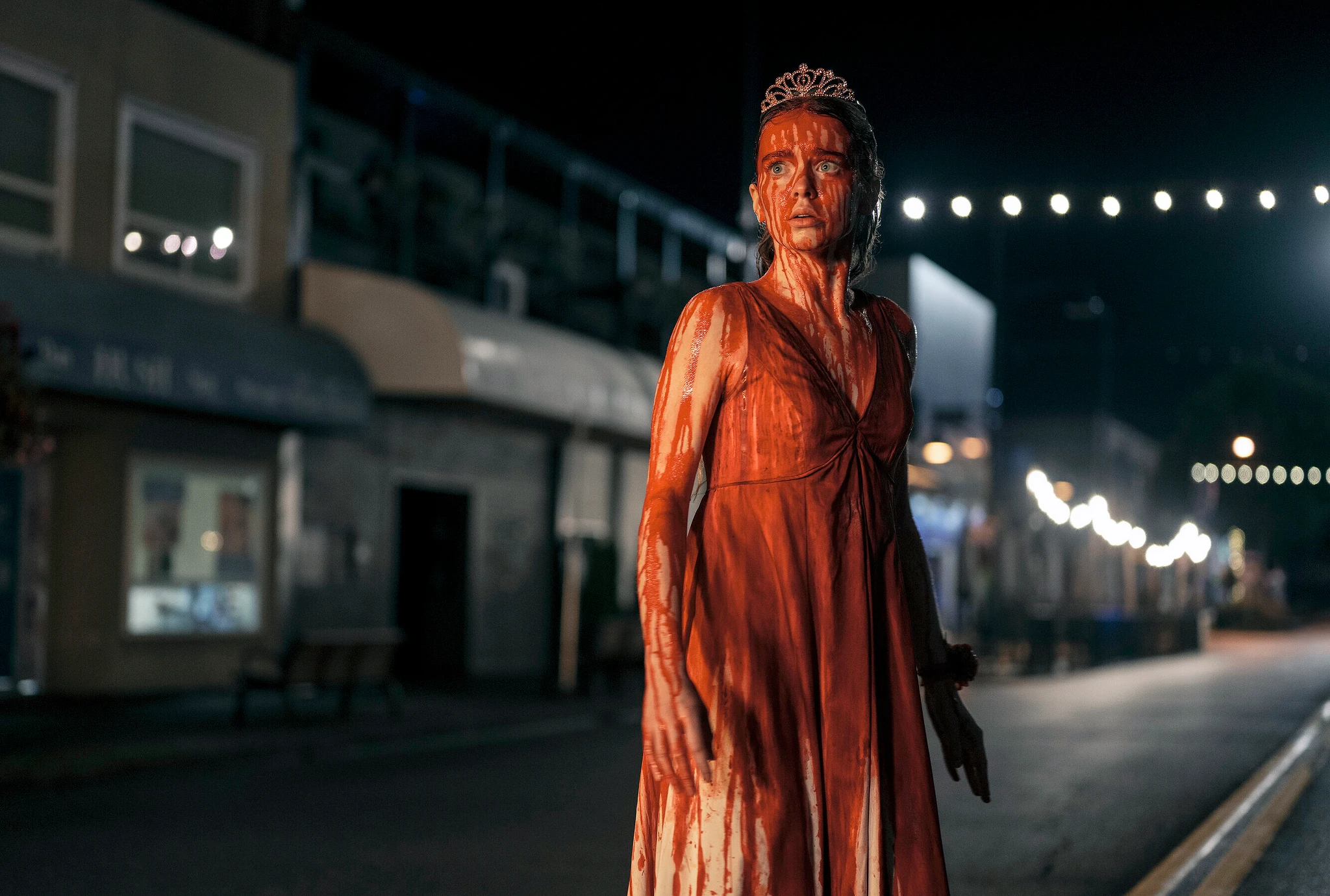 Carrie White i serien Carrie står ensam på en nattlig gata, med tiara på huvudet och en klänning dränkt i blod, med chockad blick.