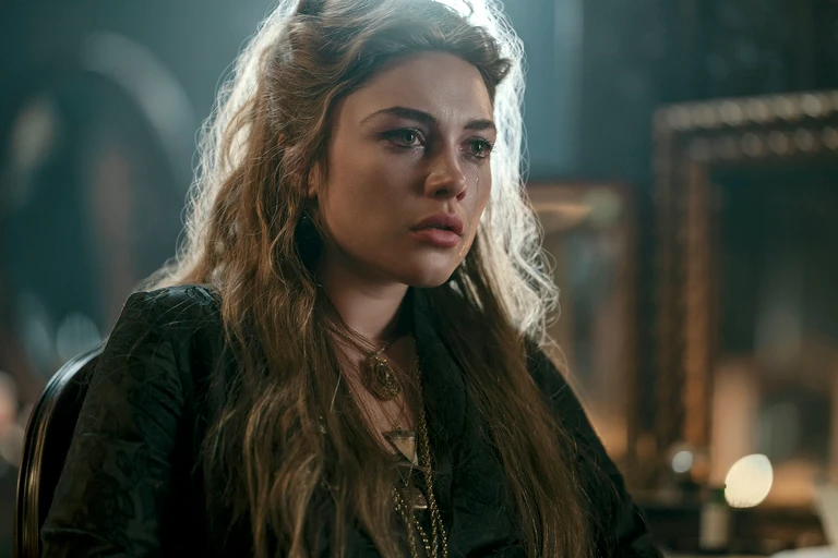 "Woman with long, wavy hair sits indoors in dim light, wearing dark clothing and a necklace, looking serious and contemplative, with blurred picture frames and a candle in the background."