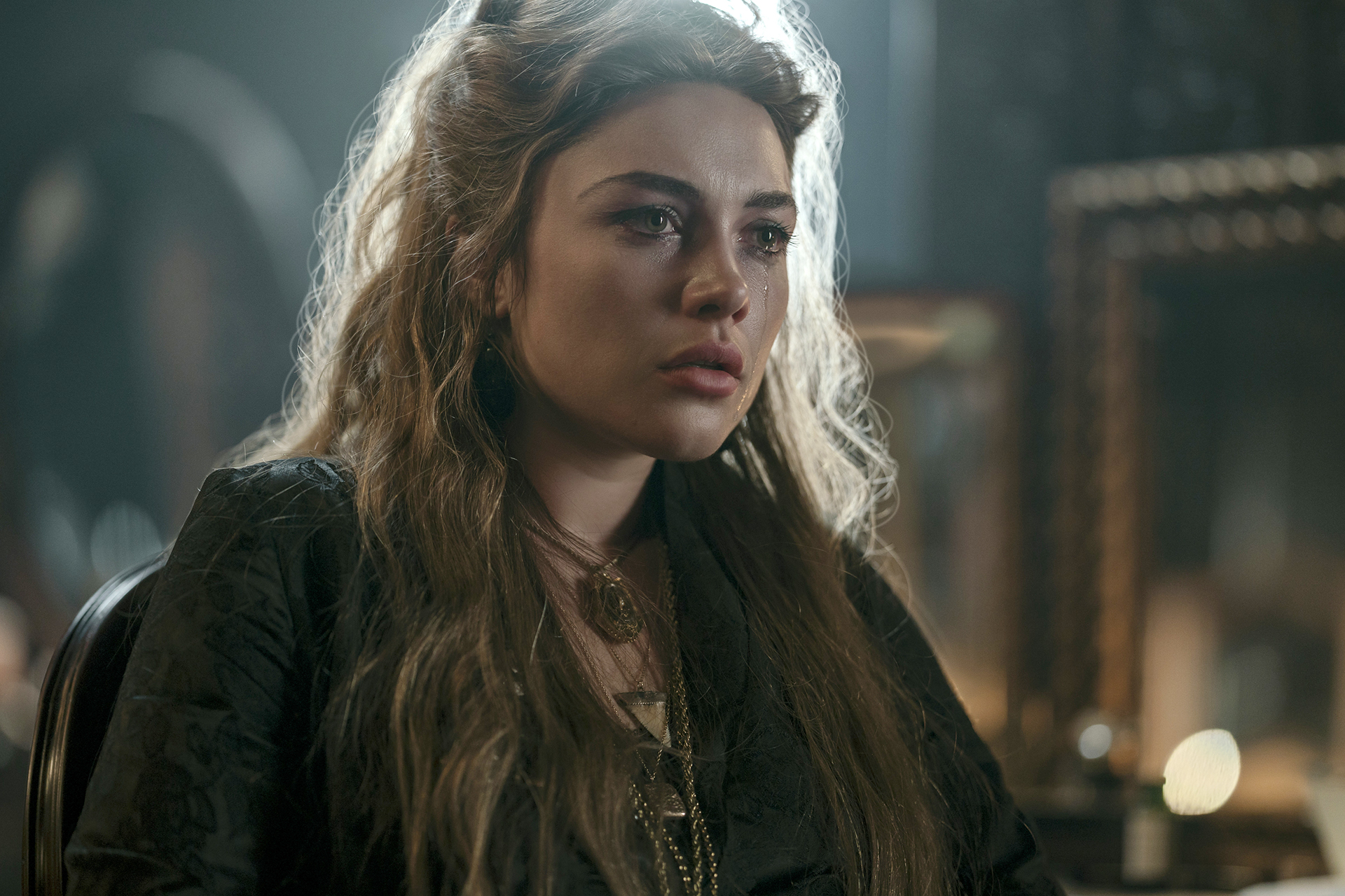"Woman with long, wavy hair sits indoors in dim light, wearing dark clothing and a necklace, looking serious and contemplative, with blurred picture frames and a candle in the background."