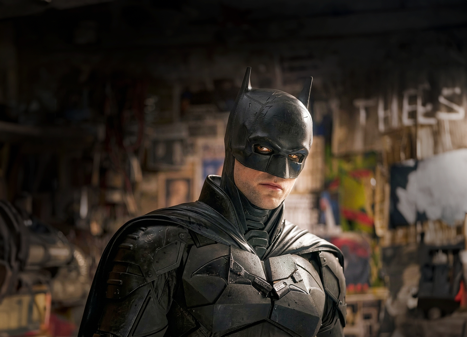 Batman i svart dräkt med mask och cape står i en belamrad, dunkel miljö och tittar allvarligt åt sidan.