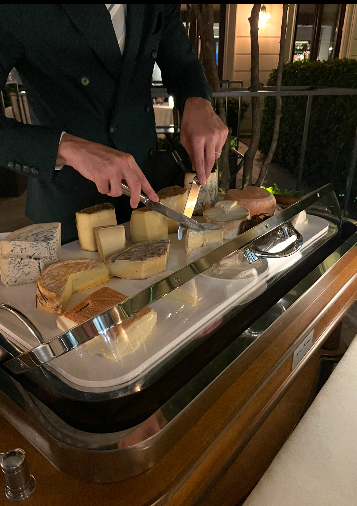 En servitör i mörk kostym skär upp ost från en ostvagn med många sorters ost, bland annat blåmögelost och lagrade ostar, på en restaurangterrass på kvällen.