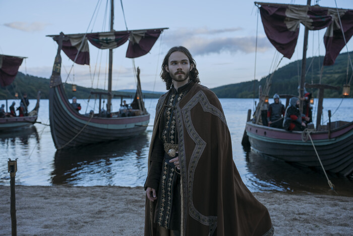 Haakon the Good, spelad av Levi Miller, står på en strand i brun, broderad kappa. Bakom honom ligger vikingaskepp med röda segel på en sjö i serien Bloodaxe.