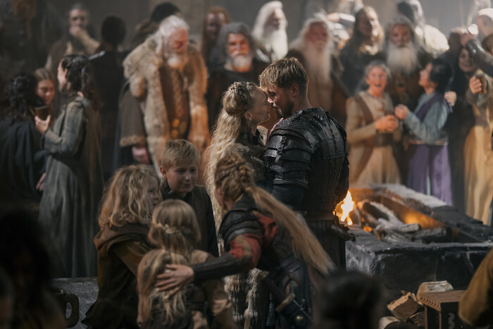 Gunhild och Erik Bloodaxe i en innerlig stund framför en eld i en fullsatt vikingahall, omgivna av barn och äldre män med långa skägg i serien Bloodaxe.