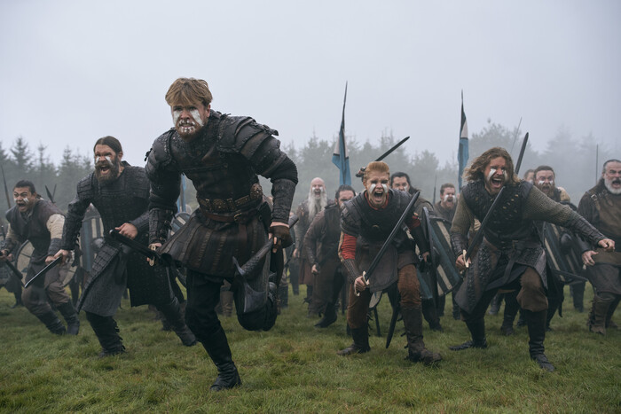 Vikingakrigare med målade ansikten rusar skrikande framåt över en gräsäng i dimma, med svärd, sköldar och blå flaggor bakom sig, i serien Bloodaxe.