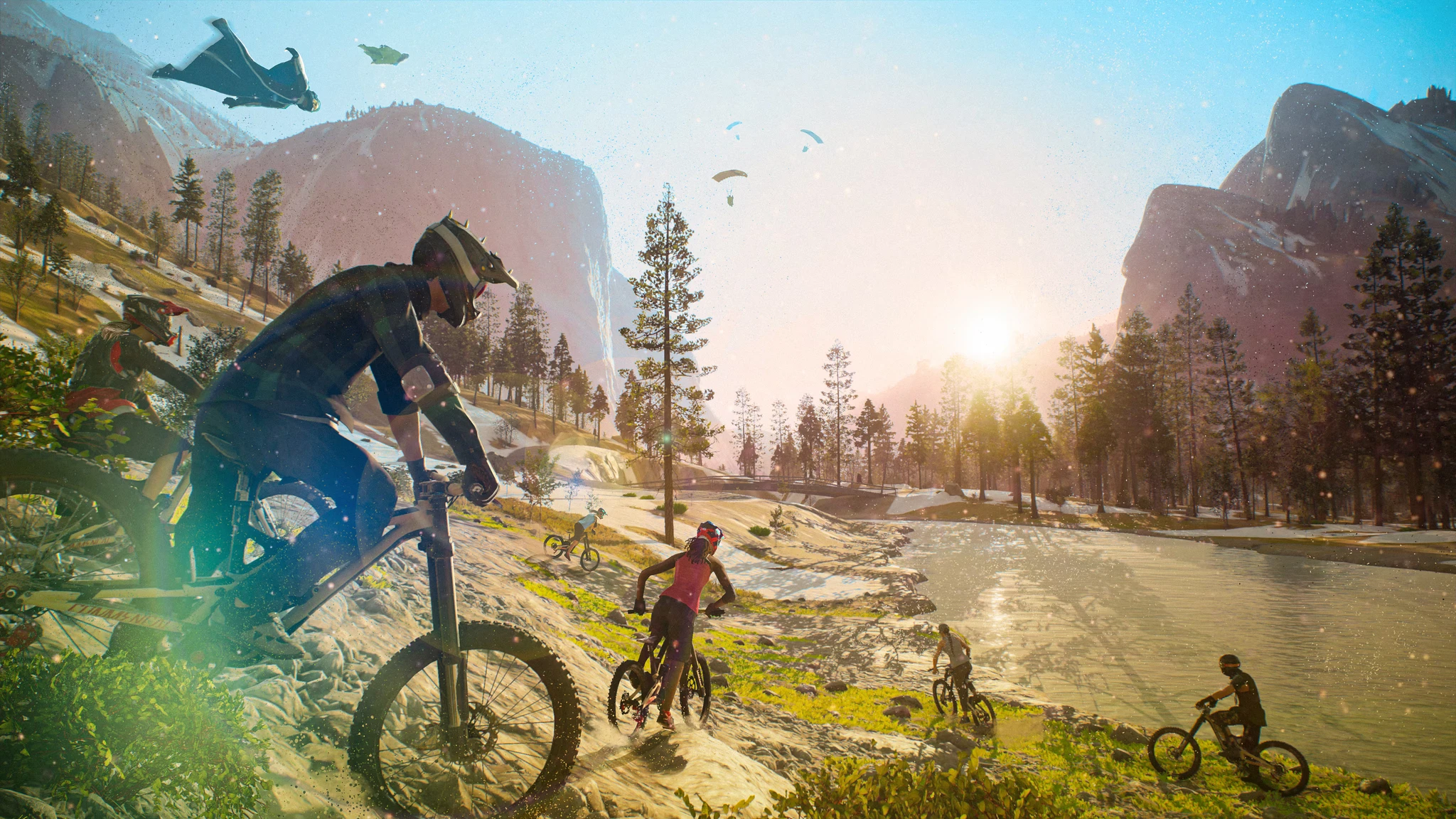 Mountainbikecyklister far nedför en stenig sluttning vid en flod i Riders Republic, med fallskärmshoppare och en wingsuit-flygare på himlen och solen lågt mellan bergen.
