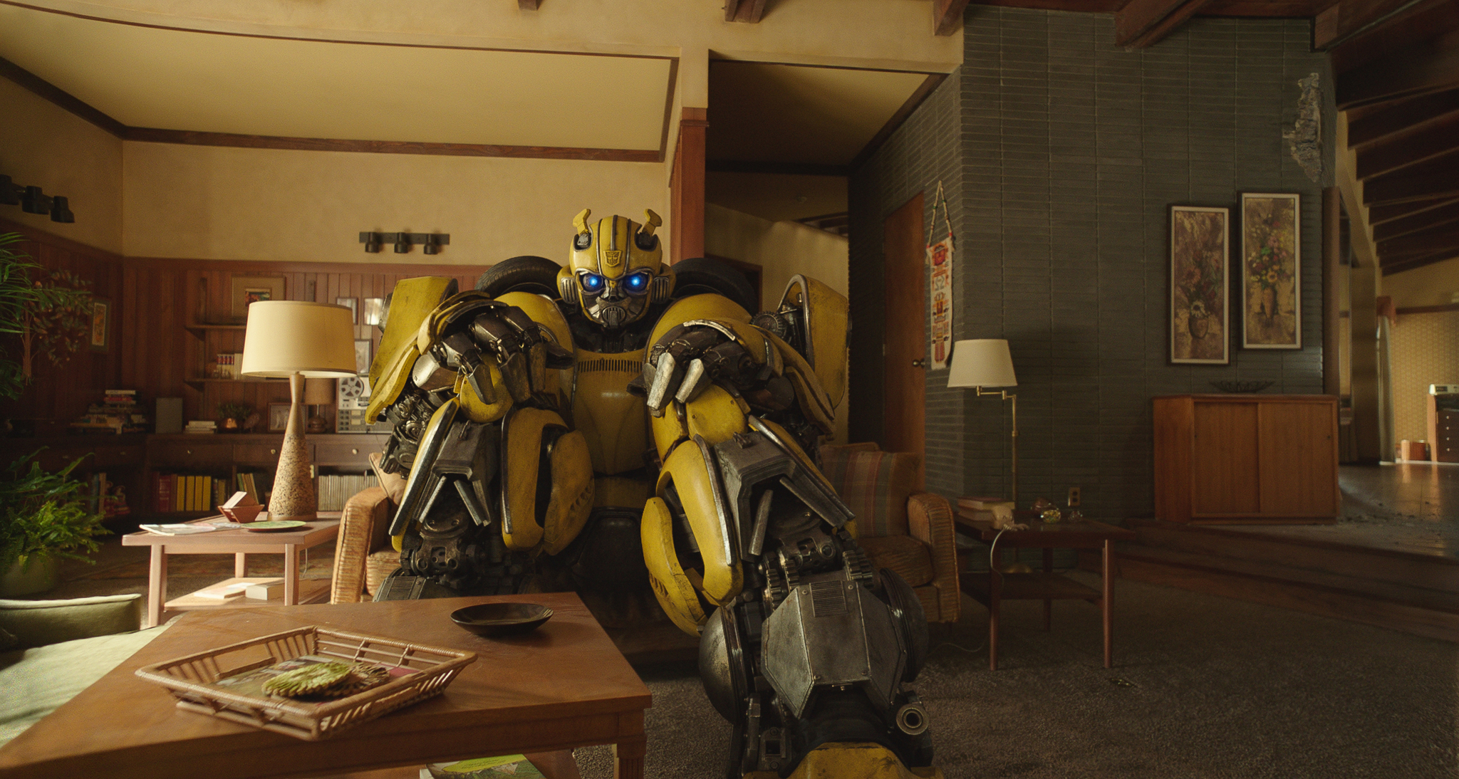 Den gula roboten Bumblebee sitter hopkrupen i ett mysigt vardagsrum i 70-talsstil med träpanel, lampor och lågt soffbord i förgrunden.