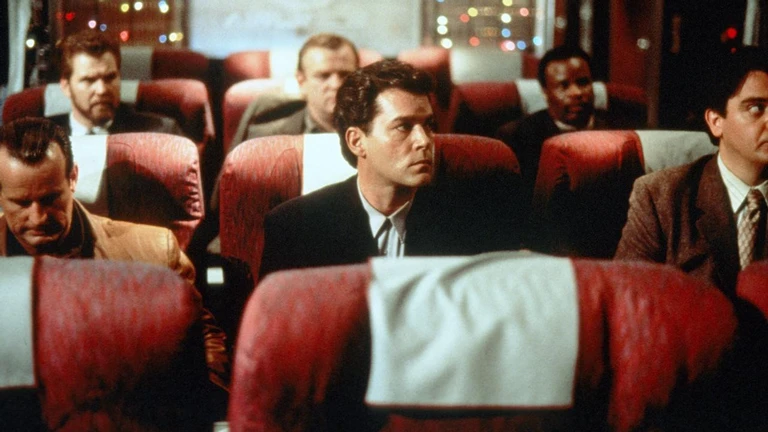 En mörkhårig man i svart kostym sitter i en röd flygplansstol och tittar spänt åt sidan, omgiven av andra passagerare i kostymer och julbelysning i fönstren.