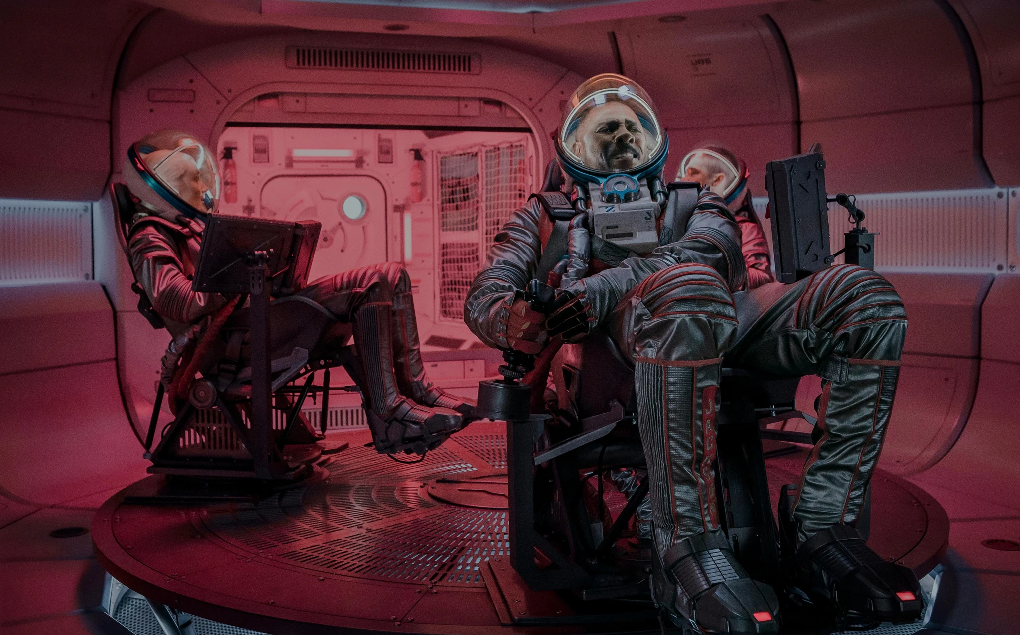 Tre astronauter i rymddräkter och hjälmar sitter i stolar i en rödbelyst rymdfarkost, med en tung maskinell dörr i bakgrunden.