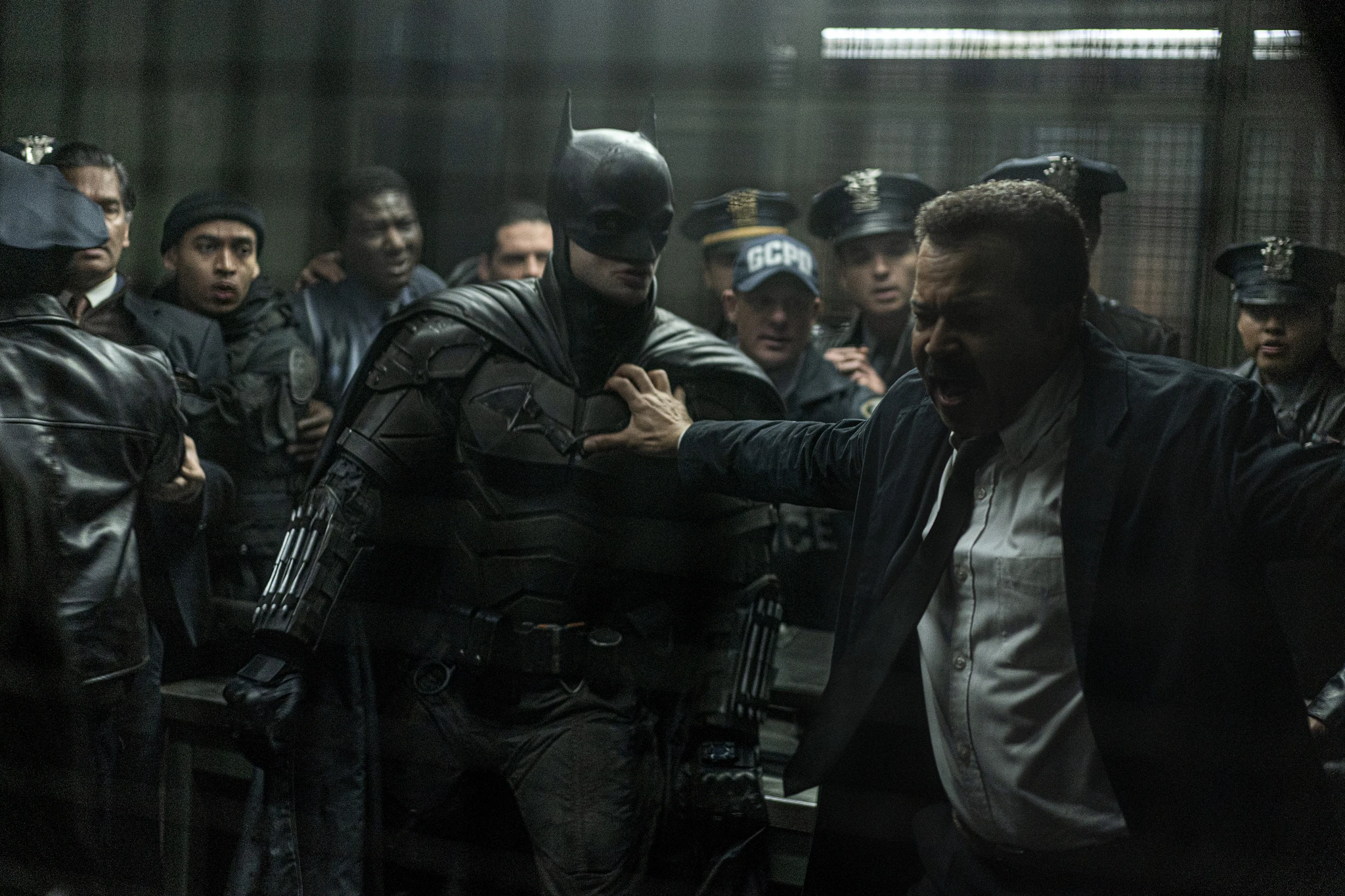 Batman i svart pansardräkt står bland poliser med GCPD-märkta mössor medan en uppretad man i kavaj sträcker ut armen och trycker mot hans bröst.