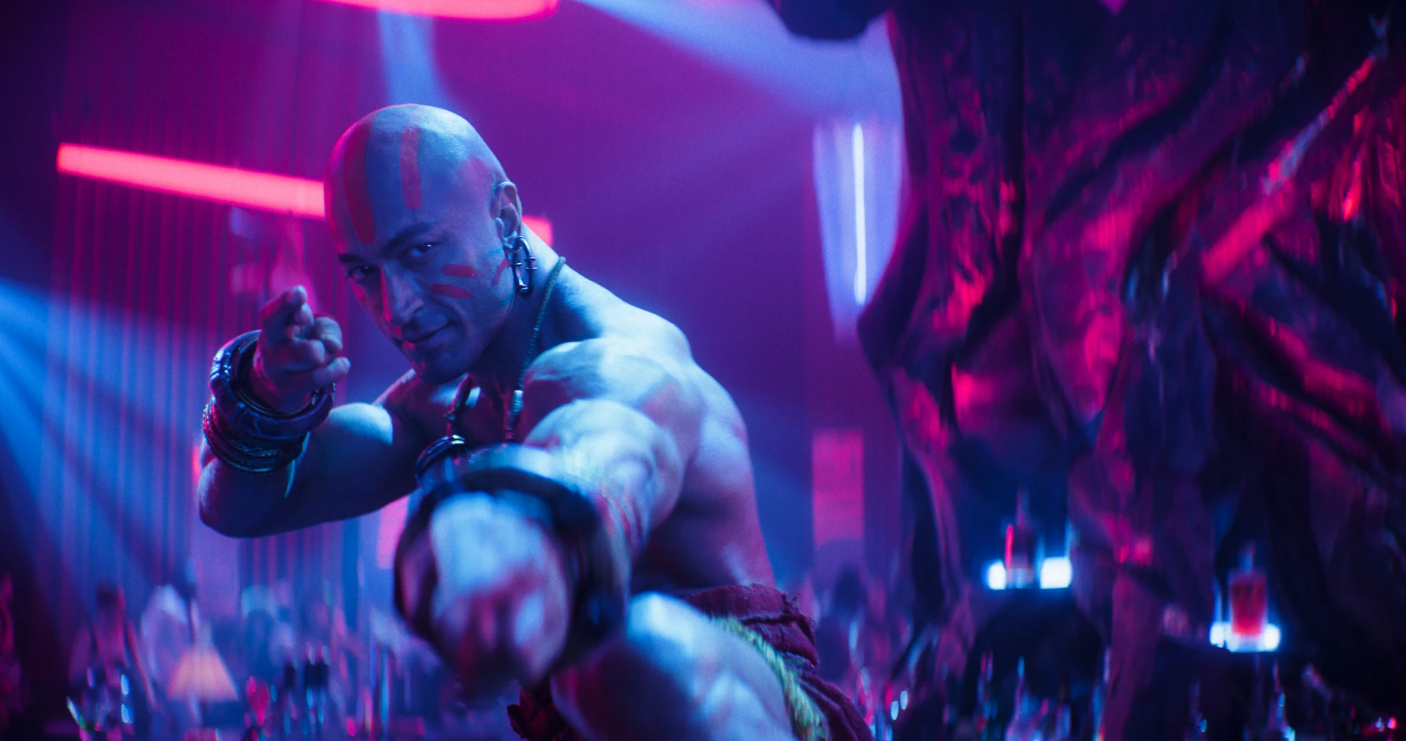 Flintskallig, bar överkropp med armband och örhänge pekar mot kameran i en neonbelyst nattklubb i rosa och blått, scen ur filmen Street Fighter.
