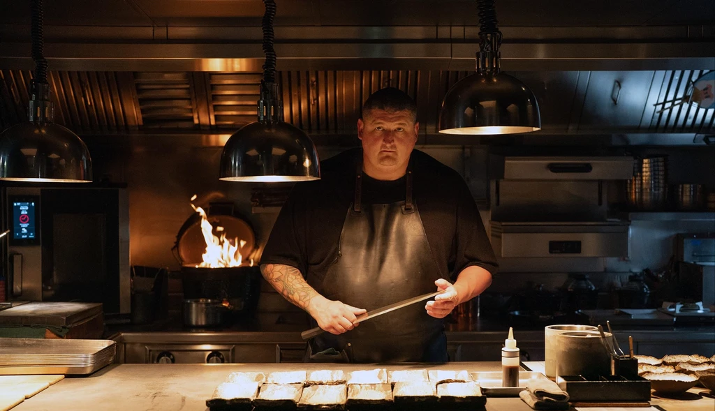 Allvarsam kock i svart förkläde står i ett mörkt restaurangkök och slipar en kniv bakom arbetsbänken, med en eld som flammar i en stekpanna bakom honom.