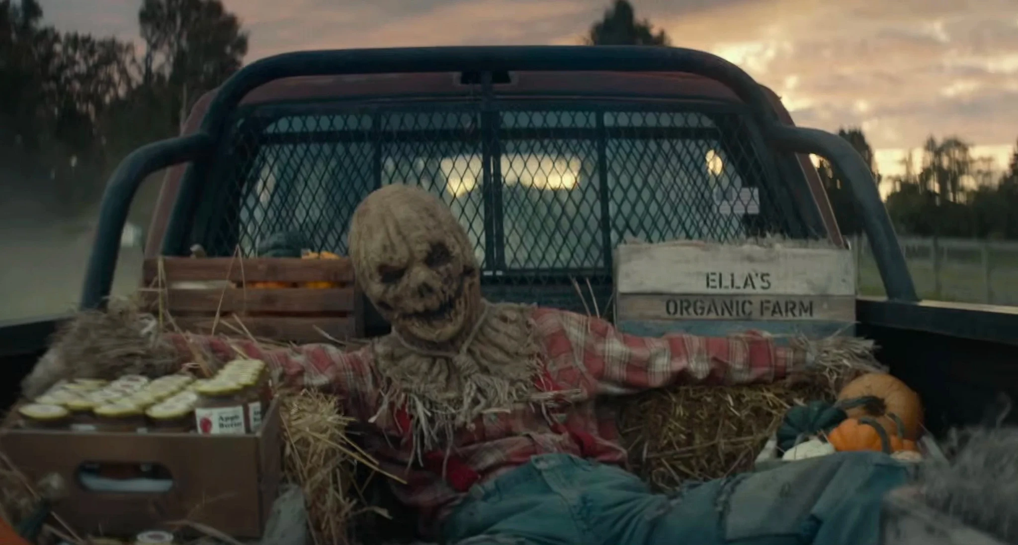 En fogliknande fågelskrämma med säckvävshuvud och rutig skjorta sitter på halmbalar på flaket av en pickup, bredvid pumpor och en skylt med texten Ella's Organic Farm.