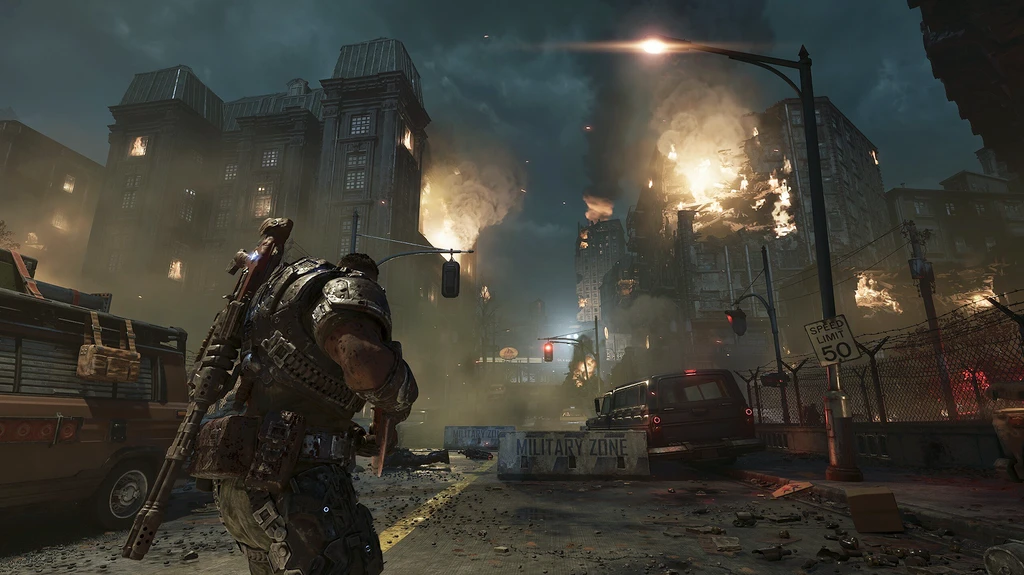 Gears of War: E-Day. En tungt bepansrad soldat med gevär på ryggen går genom en nattlig stadsgata där byggnader brinner i bakgrunden, intill ett militärspärr-staket och övergivna fordon.