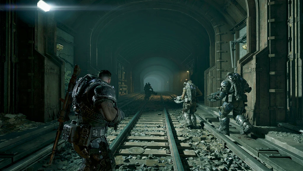 Tungt bepansrade soldater tar sig försiktigt fram längs järnvägsspåren i en mörk, förfallen tunnel i Gears of War: E-Day, med en silhuett av fiender längre bort.