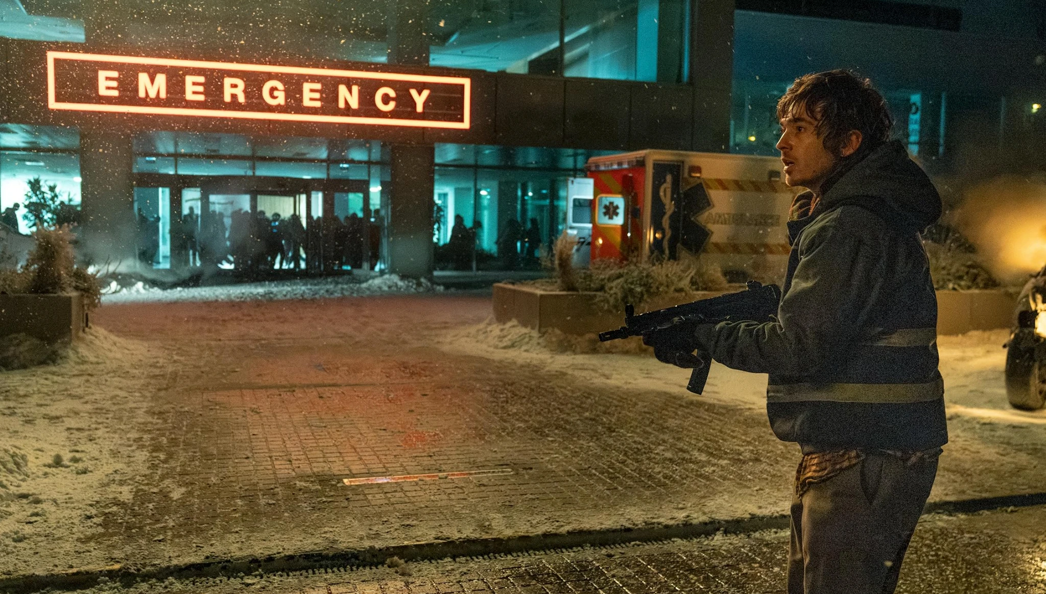 En man i jacka med reflexband håller ett automatvapen i snöfall utanför en sjukhusentré med lysande skylt "Emergency", en ambulans och en folksamling i bakgrunden.