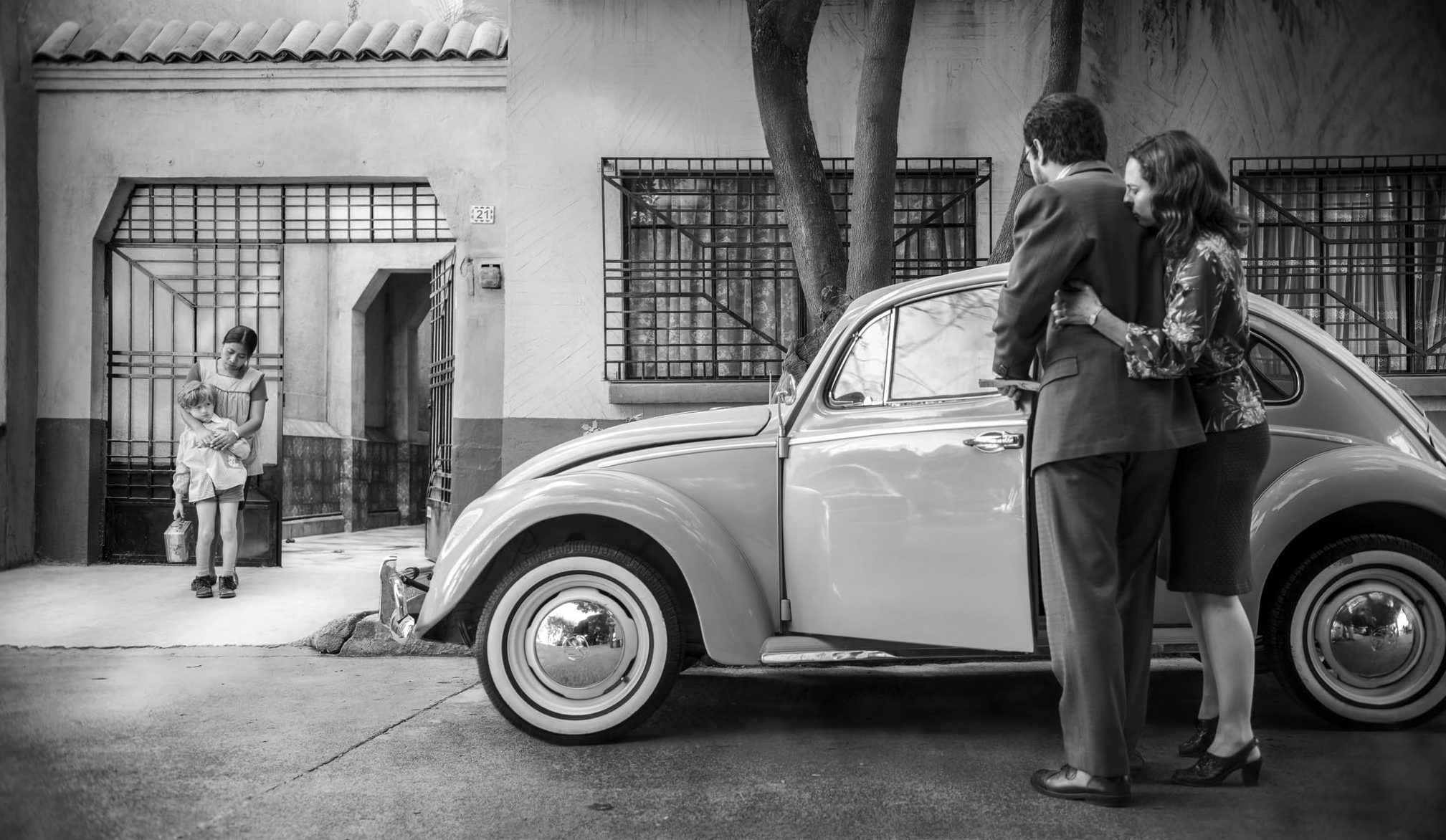 Svartvitt filmstill: ett par omfamnas intill en parkerad Volkswagen Typ 1 på en gata, medan en liten flicka och en äldre flicka står vid en grindförsedd port i bakgrunden.