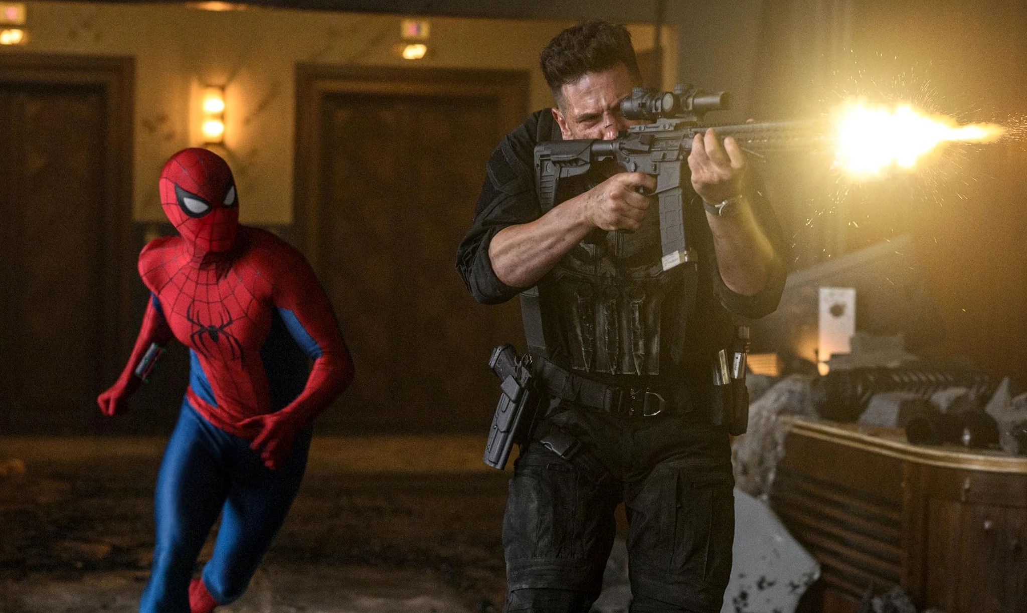 Spider-Man i rödblå dräkt springer i en dunkel hotellkorridor medan en man i svart skyddsväst skjuter med ett gevär, mynningsflammorna lyser upp rummet.