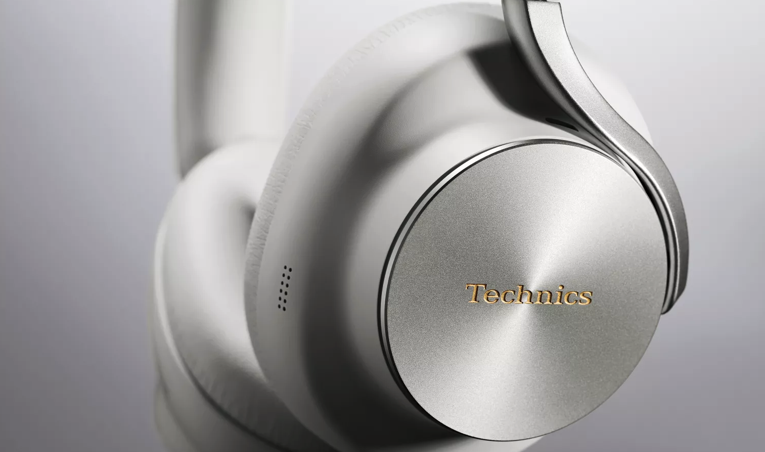 Närbild på vita hörlurar från Technics med silverfärgad öronkåpa av borstad metall och guldfärgad logotyp, mot grå bakgrund.