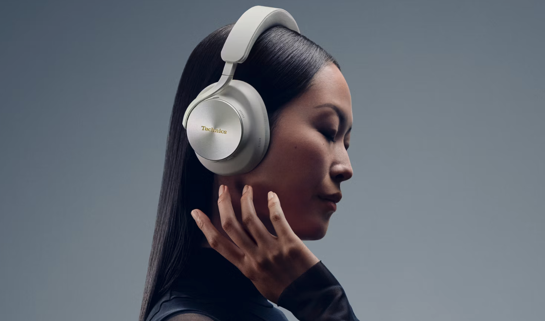 Kvinna i profil med slutna ögon och handen mot kinden bär silvervita over-ear-hörlurar från Technics, mot en grå bakgrund.