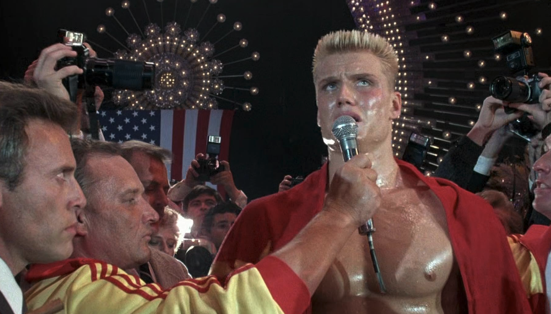 Svettig, blond och bar överkropp i röd mantel talar i en mikrofon framför en folkmassa av fotografer och en amerikansk flagga, i en scen från Rocky IV.