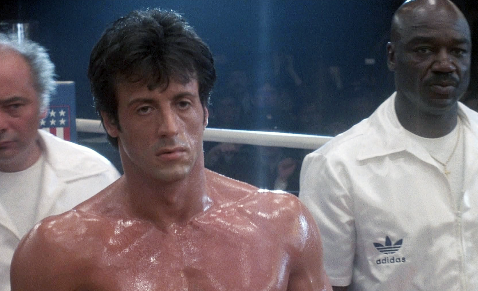 Svettig, barbröstad boxare står allvarsam i ringen i Rocky IV, flankerad av två män i vita jackor, en av dem med adidas-logga.