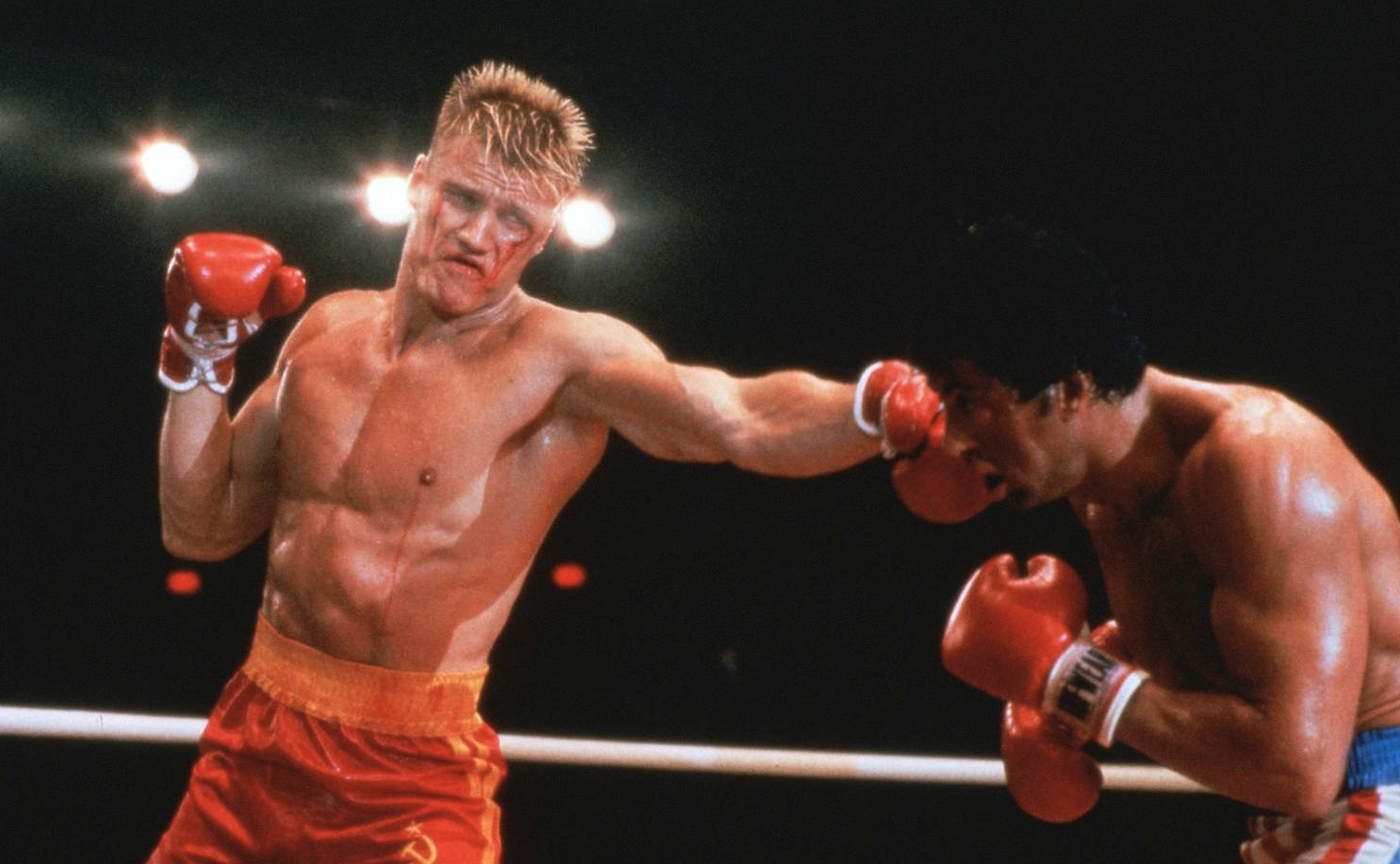 Boxare med kortklippt blont hår och röda shorts slår en rak höger mot en mörkhårig motståndare i röda handskar, mitt i en match i ringen under strålkastare.
