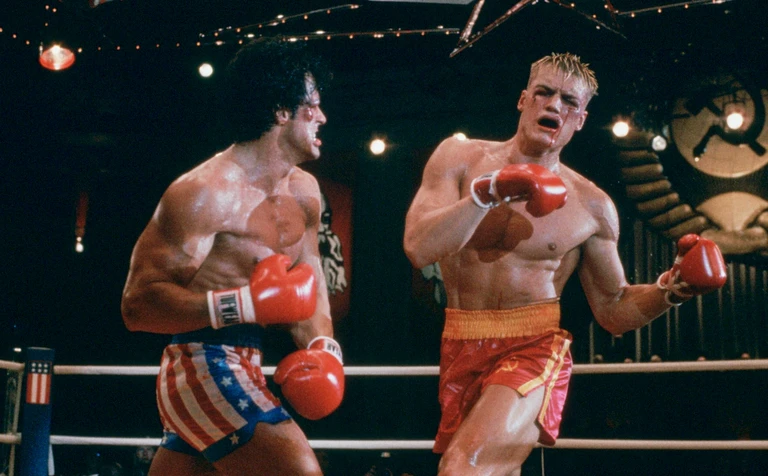 Två bröstbara boxare slåss i ringen i Rocky IV: till vänster en mörkhårig man i stjärnbannershorts, till höger en ljushårig man i röda shorts som tar emot ett slag.