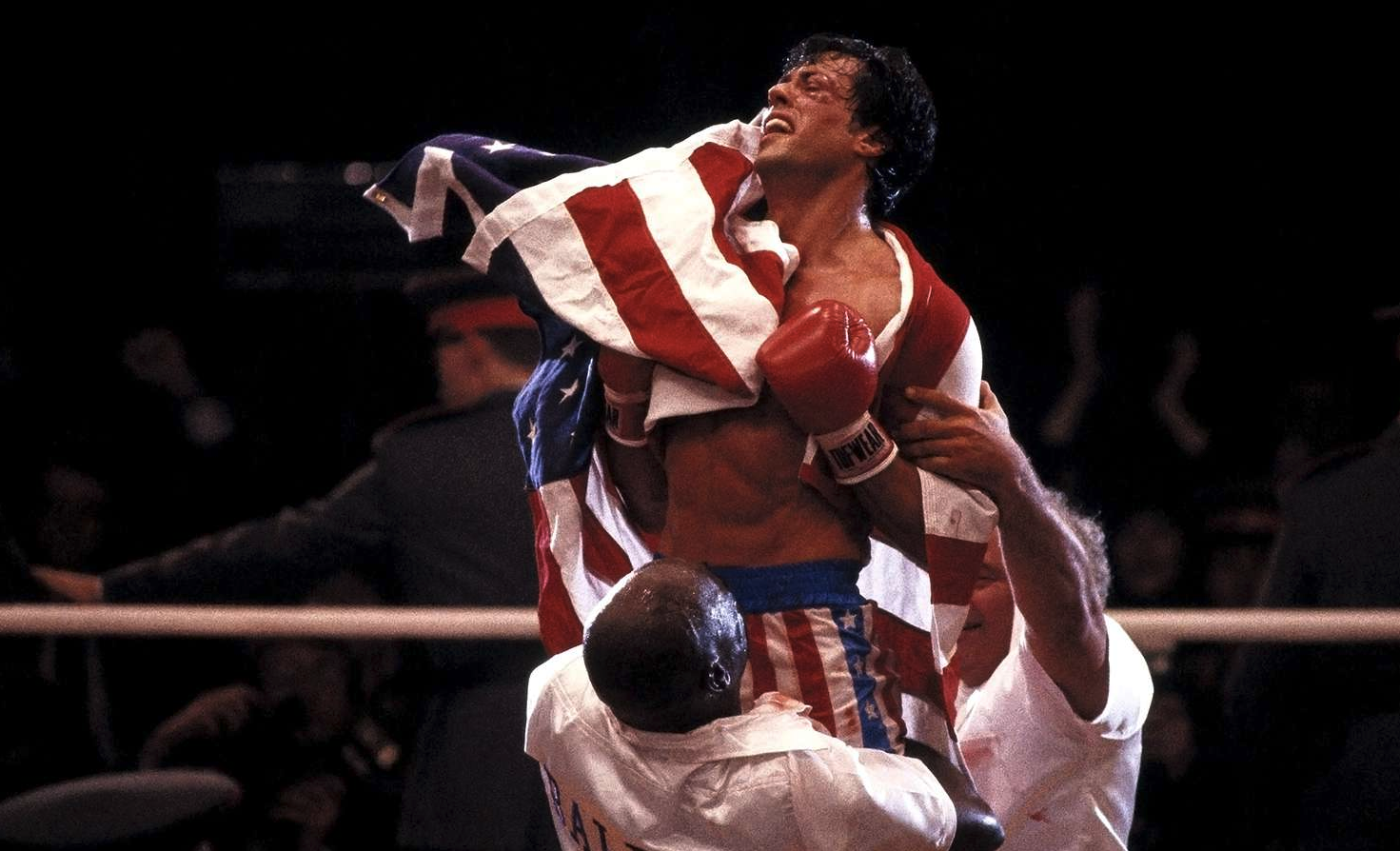 Rocky IV: en uppsvettig boxare i röda handskar och stjärnspängd shorts bärs på axlarna i ringen med en amerikansk flagga över ryggen, omgiven av jublande hjälpare.